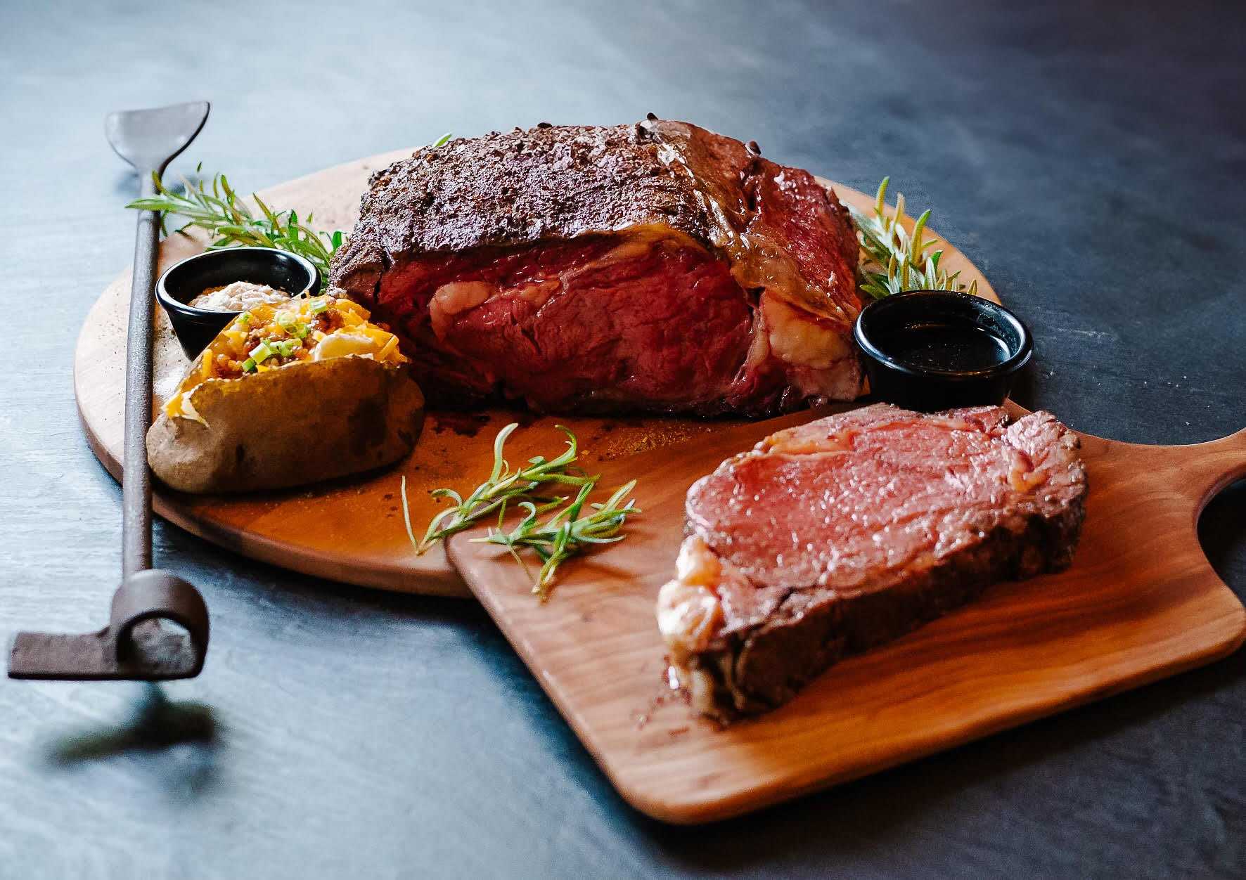 Prime rib roast on a wooden board with baked potato, herbs, and dipping sauces.