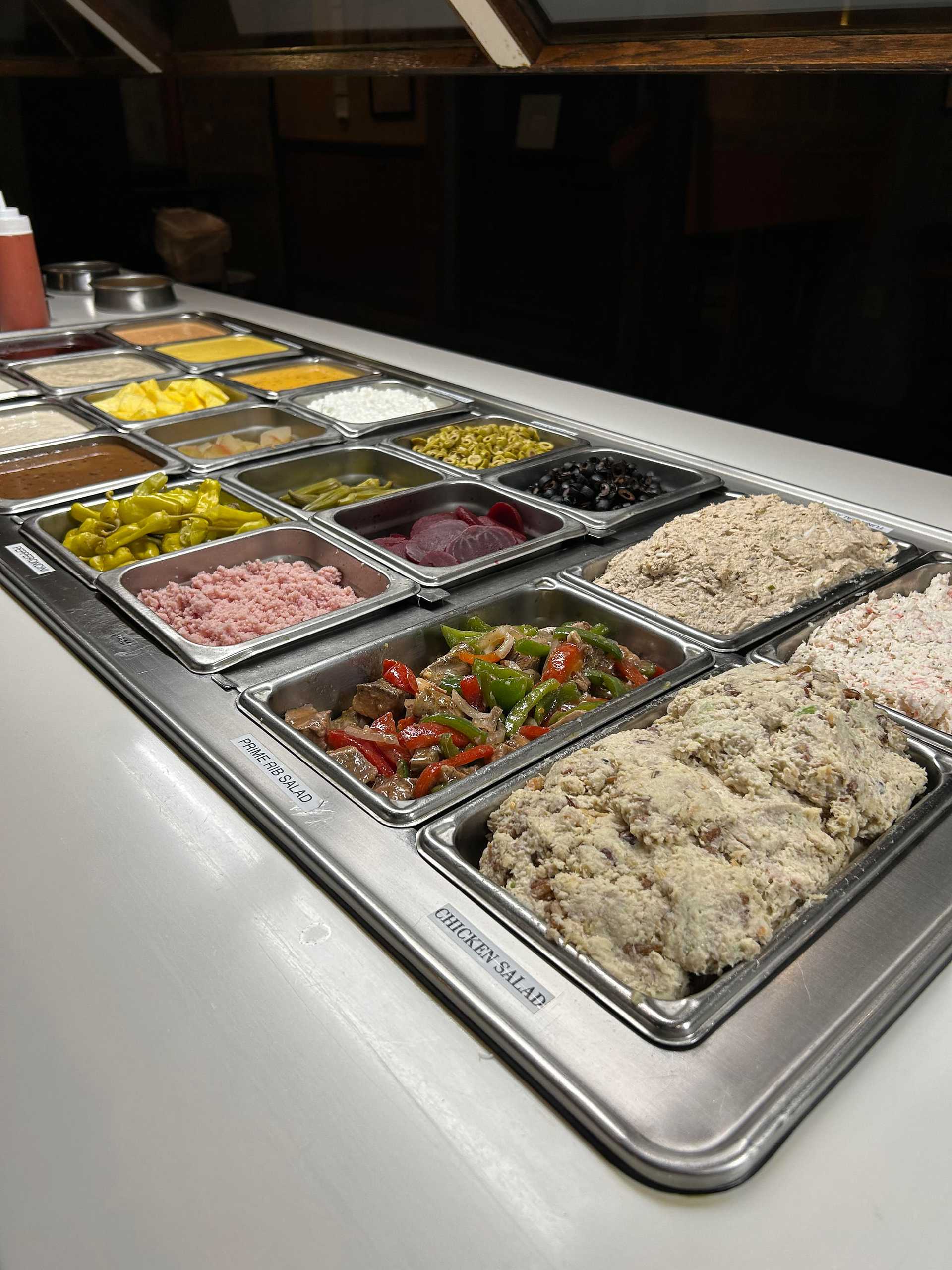 Self-serve salad bar with various fresh toppings in metal containers, including vegetables and meats.